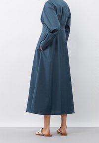 Abito lungo in teal realizzato in tessuto morbido, con maniche lunghe, corpetto aderente, tasche laterali e una gonna fluida che arriva fino al polpaccio.