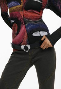 Pikendatud varrukatega pluus, millel on värvilised huulejäljed, kombineeritud saledate tumedate pükstega ja must vöö hõbedase südamekujulise pandlaga.