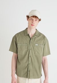 Chemise olive verte à manches courtes avec deux poches avant, fermeture à boutons et col. À associer avec une casquette beige. Texture de tissu lisse.