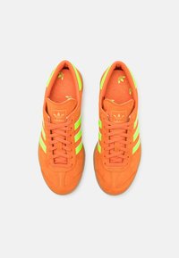 Orange suede sneakers featuring three reflective green stripes, textured insoles, and a rubber sole with a classic design.