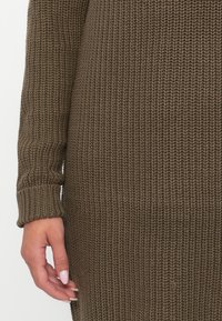 Λεπτομέρεια από καφέ πλεκτό πουλόβερ που δείχνει υφή υφάσματος, ριμπ μανσέτες και ένα χέρι τοποθετημένο στην κάτω άκρη. Μαλακό, χοντρό πλέξιμο.