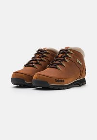 Bottes de randonnée en cuir marron avec une tige haute, semelle en caoutchouc noir et oeillets de lacet en métal. Présente un col de couleur crème et un logo "Timberland".