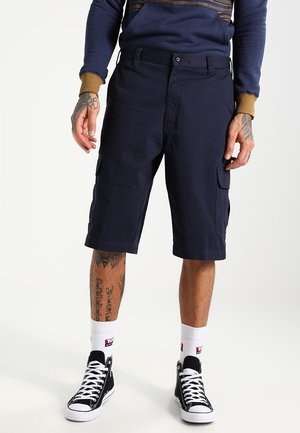 Marineblaue Cargo-Shorts mit Seitentaschen, aus Baumwollmaterial, mit umgeschlagenem Saum, kombiniert mit schwarzen High-Top-Sneakers und weißen Socken.