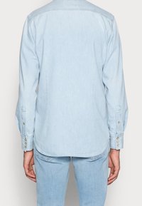Jasnoniebieska koszula dżinsowa z długimi rękawami, zapinanymi na guziki mankietami oraz zaokrąglonym dołem. Materiał ma miękką fakturę i subtelne, wyblakłe wzory.