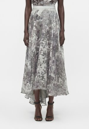 Ροδαφτή φούστα με ασύμμετρο μήκος σε γκρι και λευκό λουλουδένιο σχέδιο, φορεμένη με μαύρες ψηλοτάκουνες γόβες με λουράκι στο αστράγαλο, εμφανιζόμενη από τη μέση έως τα πόδια.