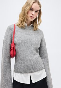 Νέα γυναίκα με κυματιστά ξανθά μαλλιά, φορώντας γκρι πουλόβερ πάνω από λευκό πουκάμισο, μαύρη φούστα και μικρή κόκκινη τσάντα ώμου σε λευκό φόντο.