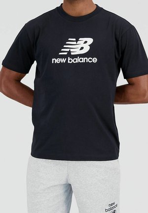 Mann trägt ein schwarzes New Balance T-Shirt mit weißem Logo und hellgraue Jogginghose mit New Balance Schriftzug am Oberschenkel.
