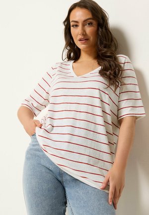 Kobieta z długimi, falującymi włosami, ubrana w białą koszulkę z czerwonymi paskami i jasnoniebieskie dżinsy, pozująca z jedną ręką w kieszeni na tle białej ściany.