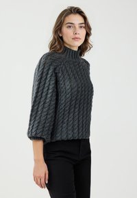 Maglione grigio a maglia con colletto alto, caratterizzato da un motivo testurizzato e maniche a tre quarti. Indossato con pantaloni neri.