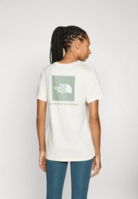 Camiseta de algodón en blanco roto con un gráfico cuadrado verde que presenta el logo de "The North Face" y el texto "NUNCA DEJES DE EXPLORAR" debajo. Mangas cortas.