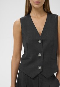 Chaleco entallado gris oscuro con un escote en V profundo, cuatro botones y dos bolsillos frontales. Tejido suave con un ajuste estructurado y dobladillo en punta.