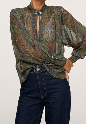 Transparante blouse met lange mouwen en groen paisleypatroon, keyhole-halslijn met metalen oogjes, ingestopt in een high waist donkerblauwe spijkerbroek.