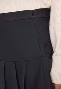 Musta laskostettu hame, jossa on sileä, teksturoitu kangas ja sivuvetoketju. Yhdistetty kermaisen väriseen palmikkoneuleeseen.