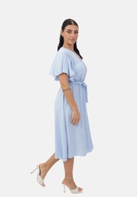 Vestido azul claro hasta la rodilla con mangas cortas drapeadas, diseño envolvente y cinturón de tela. La modelo lleva mules con tacones dorados. Textura suave.