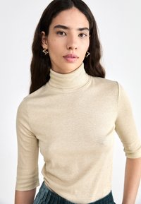 Beige ribbed poolokauluksella, jossa on kolmanneksen pituiset hihat, valmistettu pehmeästä, hieman kiiltävästä materiaalista, yhdistettynä teksturoituun vihreään hameeseen.