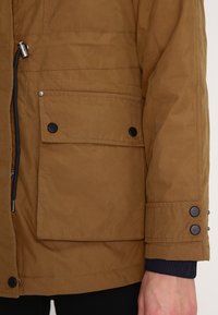 Brun parkasjacka med dragkedja, stor framficka och svarta knappdetaljer. Materialet har en mjuk textur med söm-detaljer.