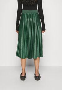 Žena oblečená v lesklé zelené skládací sukni střední délky, černém topu s dlouhým rukávem a černých robustních mokasínech, stojící čelem k bílému pozadí.