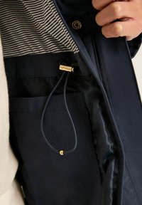 Jūras zilās jakas iekša ar svītrotu rakstu, kurai ir zelta detaļa un regulējama aukla, izceļot gludu auduma tekstūru.