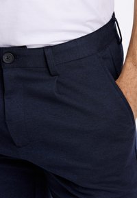 Marinblå byxor av strukturerat tyg med knappstängning, framfickor och en slimmad passform. Diskret melerad mönster.