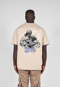 Beige short-sleeve t-shirt featuring a graphic print of two figures on the back with blue accents and a textured finish.