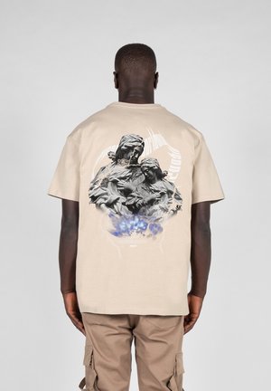 Beige kortærmet t-shirt med grafisk print af to figurer på ryggen med blå accenter og en tekstureret finish.