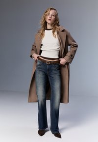 Brun överrock över en vit ribbad topp, i kombination med bleka blå bootcut-jeans och ett brunt bälte, accentuerat av mörkbruna klackar.
