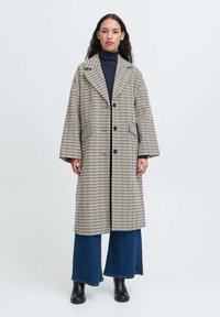 Langternet frakke i beige og marineblå med et kæbeformet krave, dobbelte knapper og sidelommer, båret over vide denimbukser.