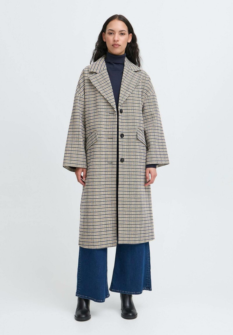 Langternet frakke i beige og marineblå med et kæbeformet krave, dobbelte knapper og sidelommer, båret over vide denimbukser.