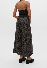 Pantalons à jambes larges à motifs noirs avec des designs circulaires dorés. Dotés d'une taille haute et d'une silhouette ample et fluide. À associer avec des sandales à plateforme noires.