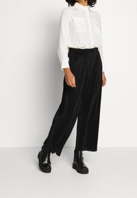 Pantalon noir à jambes larges en tissu texturé avec une ceinture élastique, associé à une chemise blanche à manches longues boutonnée, dotée de deux poches poitrine.
