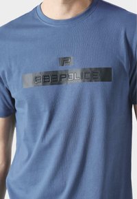 Sininen puuvillapaita, jossa on kiiltävä musta logo ja teksti "883POLICE" edessä, klassisella pyöreällä kaula-aukkodesignilla.