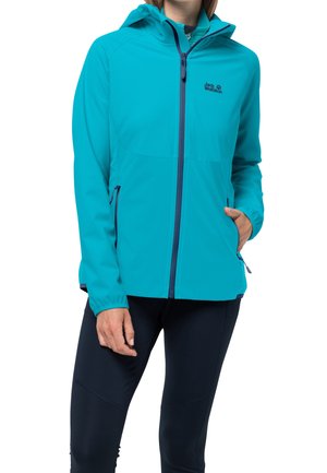 Femme portant une veste de plein air bleu vif à capuche avec poches zippées et pantalon noir, debout avec une main dans la poche.