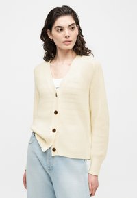Νεαρή γυναίκα με σγουρά μαλλιά μήκους μέχρι τους ώμους, φορούν μια κρεμ πλεκτή ζακέτα με μεγάλα καφέ κουμπιά και ανοιχτό μπλε τζιν.