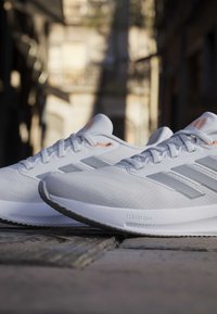 Chaussures de running Adidas blanches avec des rayures gris clair et des semelles Cloudfoam posées sur un pavé sous une lumière douce du soleil.