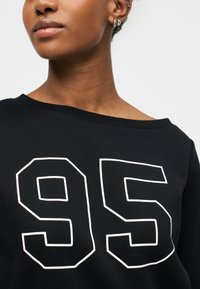 Μαύρο φούτερ με φαρδύ λαιμόκοψη, με μεγάλο λευκό τυπωμένο "95" στο μπροστινό μέρος. Λείος υφή και άνετη εφαρμογή.