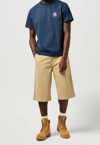 Camiseta de algodón de color navy con un pequeño logo, pantalones cortos de lona color caqui hasta la rodilla y botas marrones con cordones, con detalles en negro y suelas de goma.