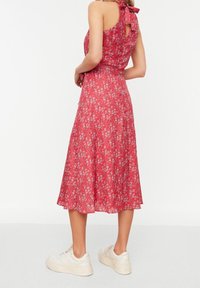 Kvinna klädd i en röd blommig midiklänning med halterneck som är knuten i ryggen, kombinerad med vita platåskor, stående mot en neutral bakgrund.