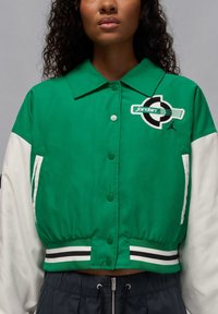 Zaļš jakas vēzis ar baltiem piedurknēm, pogu priekšpusi, melnbaltu svītru apakšmalu un izšūtu logotipu kreisajā krūtī.