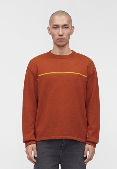 Homme au crâne rasé portant un pull orange rouille avec une fine ligne jaune horizontale sur la poitrine, debout devant un fond clair uni.
