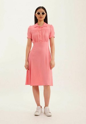 Robe rose à manches courtes avec un col, des boutons décoratifs et une jupe évasée. Portée avec des baskets blanches et des lunettes de soleil rondes surdimensionnées.