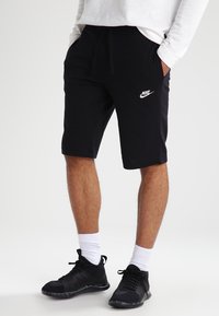 Svarta Nike-shorts med elastisk midja, prydda med en vit logotyp. I kombination med svarta träningsskor och vita strumpor. Tyget är en blandning av bomull.