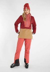 Rode en tan buitenjas met een ritszak aan de voorkant, gecombineerd met bijpassende roze broek en zwarte skischoenen. Bevat een gebreid muts.