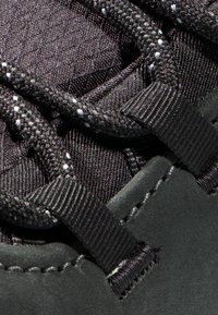 Detail eines schwarzen Sportschuhs mit strukturiertem Gewebe, schwarzen Schnürsenkeln mit silbernen Akzenten und glattem, wildlederähnlichem Material im unteren Bereich.