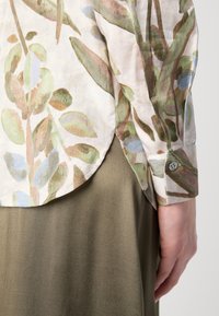 Camicia leggera con motivo floreale realizzata in un tessuto morbido; presenta motivi vegetali verdi e blu con orlo curvo e dettagli di polsino con bottoni.