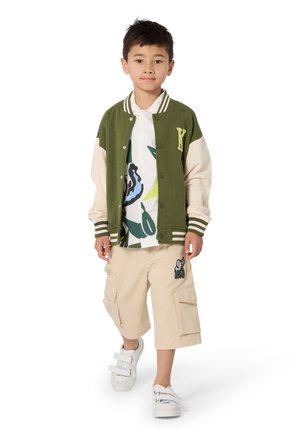 Veste universitaire verte et crème, chemise blanche graphique avec motifs de feuilles, shorts cargo beige avec poches, et baskets blanches à bride.