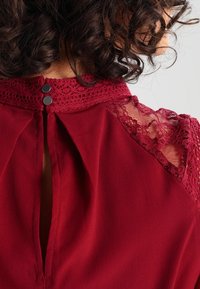 Blouse rouge avec un col montant, des accents en dentelle sur les épaules, un dos en trou de serrure et une fermeture à double bouton. Tissu doux et texturé.