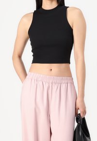 Černý žebrovaný crop top s vysokým krkem, doplněný o růžové kalhoty s elastickým pasem. Modelka drží černou tašku, čímž předvádí hladkou texturu látky.