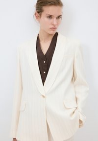 Vit kavaj med smala ränder, med notch-lapel, en enda framknapp, två fickor och skräddarsydd passform, bärs över en brun topp med knapprad.