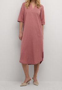 Rosenfarbendes Leinenkleid mit V-Ausschnitt, kurzen Ärmeln und seitlichem Schlitz. Verfügt über eine einzelne Tasche auf der Vorderseite für zusätzlichen Detail.