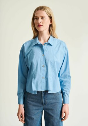 Femme blonde portant une chemise bleu clair boutonnée et un jean bleu, debout devant un fond clair uni, regardant légèrement à droite.